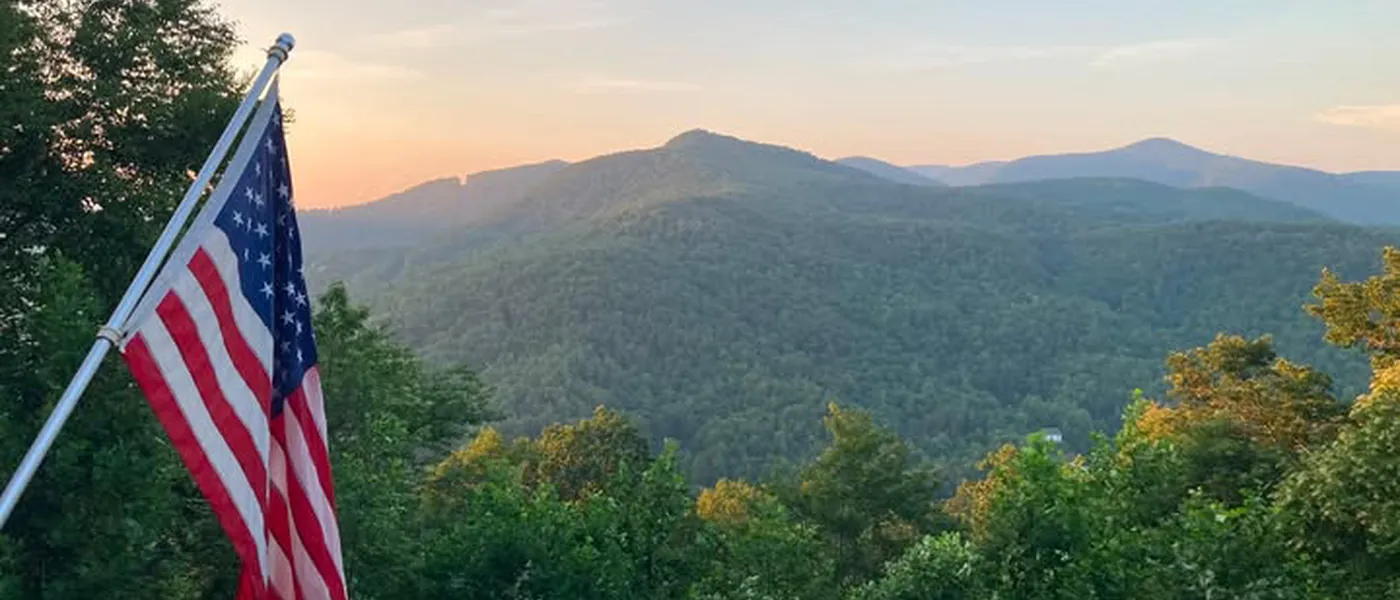The mountain view from a cleared property at sunset, ridgelines visible past the treeline with an American flag on the deck.